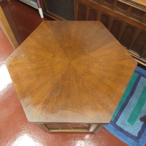 May include: A hexagonal wooden side table with a rich, warm brown finish. The tabletop features a starburst pattern, and the table has a single drawer. The table is a classic piece of furniture.