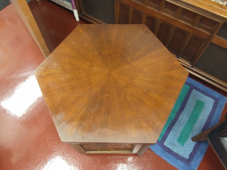 May include: A hexagonal wooden side table with a rich, warm brown finish. The tabletop features a starburst pattern, and the table has a single drawer. The table is a classic piece of furniture.