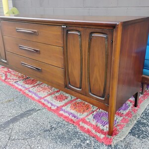 May include: A long, rectangular wooden sideboard featuring a rich brown finish. The piece includes three central horizontal drawers with dark, elongated handles, flanked by two cabinet doors on either side, each with a recessed, vertical panel design.