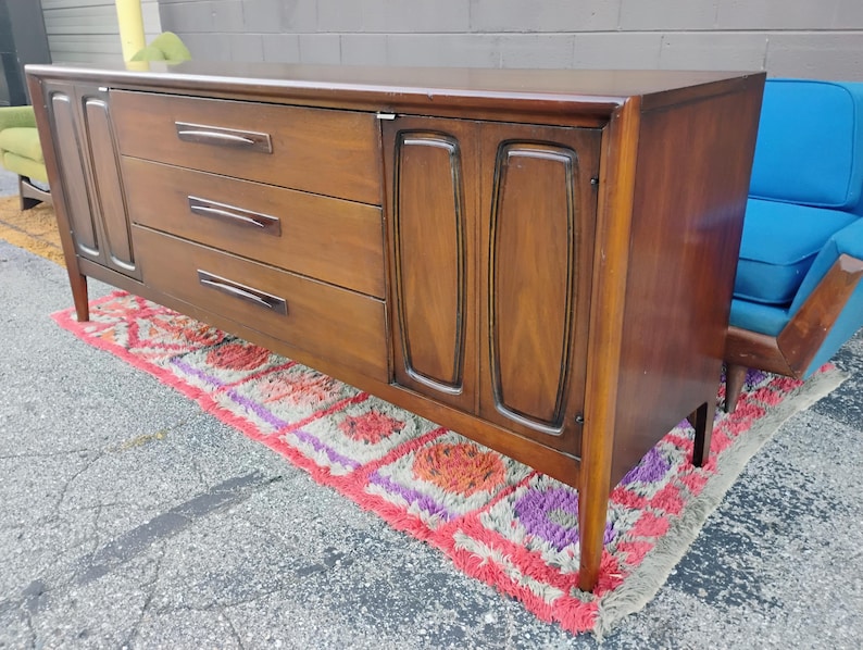 May include: A long, rectangular wooden sideboard featuring a rich brown finish. The piece includes three central horizontal drawers with dark, elongated handles, flanked by two cabinet doors on either side, each with a recessed, vertical panel design.