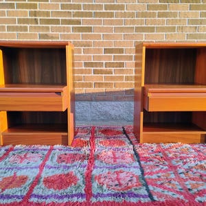 May include: Two matching wooden nightstands with a warm, reddish-brown finish. Each nightstand features a drawer and an open shelf below, with a rectangular top and a recessed back. The nightstands are placed on a colorful rug.