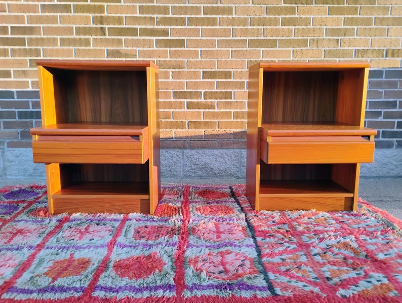 May include: Two matching wooden nightstands with a warm, reddish-brown finish. Each nightstand features a drawer and an open shelf below, with a rectangular top and a recessed back. The nightstands are placed on a colorful rug.