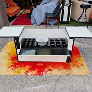 May include: A black and white bar cart with two fold-down side tables. The cart has a built-in ice bucket and a glass shelf. The cart is sitting on a colorful rug.
