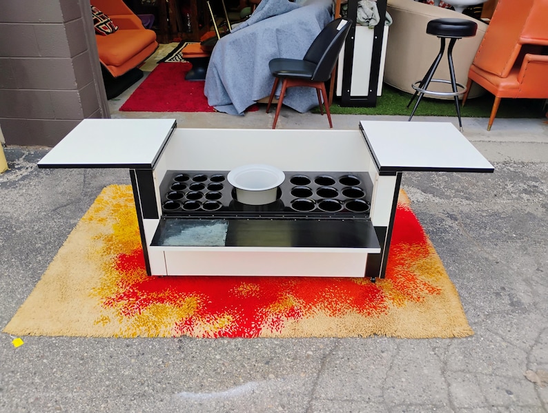 May include: A black and white bar cart with two fold-down side tables. The cart has a built-in ice bucket and a glass shelf. The cart is sitting on a colorful rug.