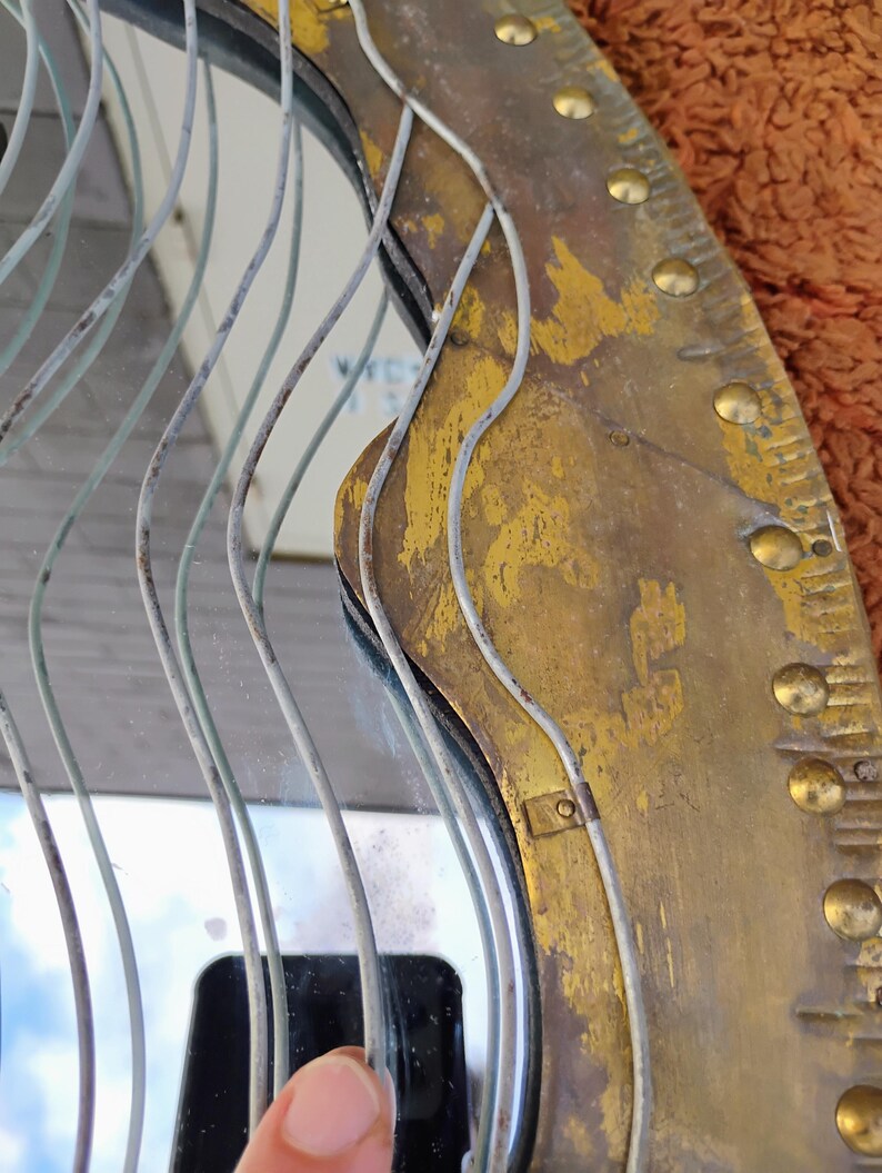 K&ouml;nnte beinhalten: Ein runder Vintage-Spiegel mit einem Messingrahmen. Der Rahmen hat ein abgenutztes, gealtertes Aussehen mit goldener Oberfl&auml;che und dekorativen Nieten. D&uuml;nne, wellenf&ouml;rmige Metalldr&auml;hte sind vor der Spiegeloberfl&auml;che angeordnet.