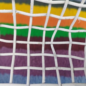 May include: A white wire mesh grid with a square pattern overlaying a background of horizontal stripes in yellow, orange, green, dark red, purple, and blue. The white metal wires are woven together to form a consistent, repeating open square structure.