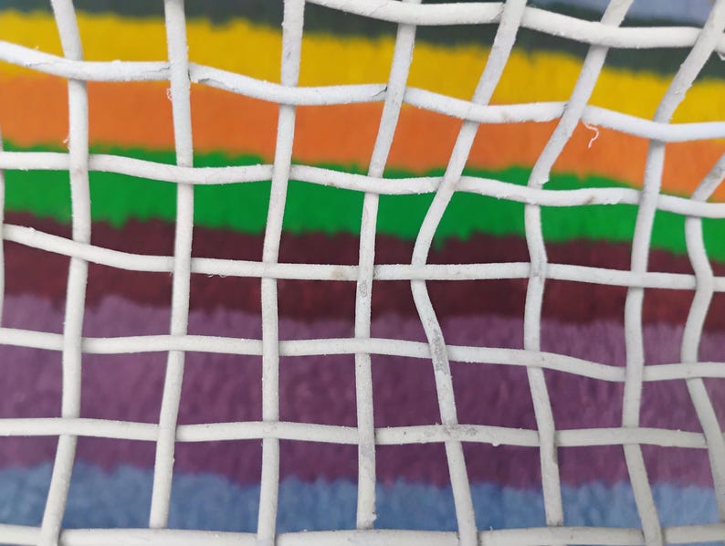 May include: A white wire mesh grid with a square pattern overlaying a background of horizontal stripes in yellow, orange, green, dark red, purple, and blue. The white metal wires are woven together to form a consistent, repeating open square structure.