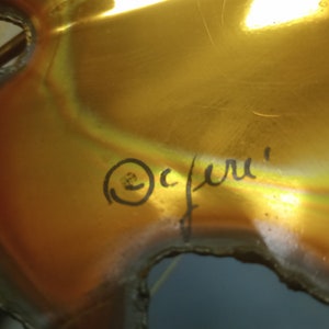 Puede incluir: Una escultura de metal de color dorado con una superficie rugosa y texturizada. La escultura est&aacute; firmada "Ocfuru" en tinta negra.