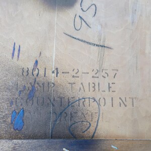 May include: Close-up of a light brown wooden table top with a label that reads "3014-22-257  DROP TABLE COUNTERPOINT  PRES", with a faded blue stain on the left side.