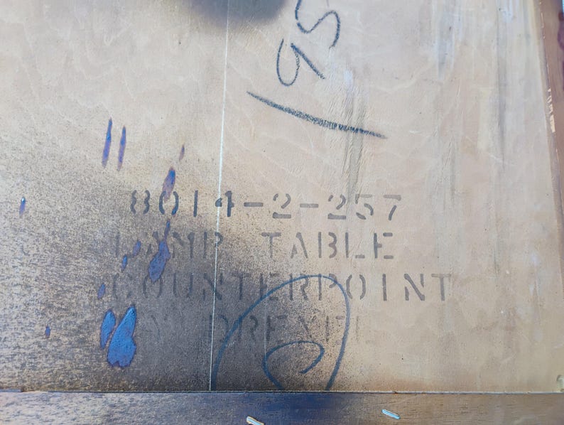 May include: Close-up of a light brown wooden table top with a label that reads "3014-22-257  DROP TABLE COUNTERPOINT  PRES", with a faded blue stain on the left side.