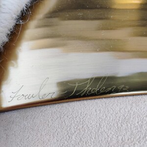 May include: A close-up of a brass-coloured rectangular object with the handwritten inscription "Fowler - Tholen 93". The object has a reflective surface with light brown and gold tones. The object is set against a white and beige textured background.