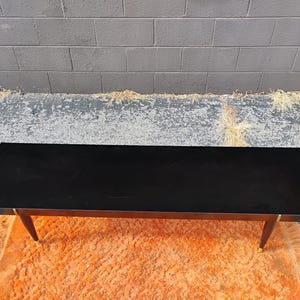 May include: A rectangular coffee table with a smooth, solid black top surface. The table features four tapered wooden legs that angle outward, each finished with a metallic gold-toned cap at the base, providing a stable and elevated structure.