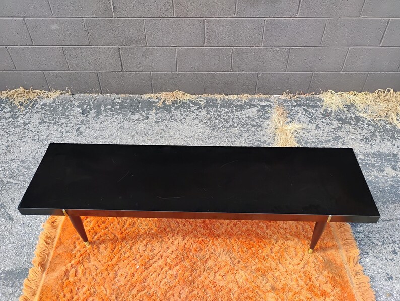 May include: A rectangular coffee table with a smooth, solid black top surface. The table features four tapered wooden legs that angle outward, each finished with a metallic gold-toned cap at the base, providing a stable and elevated structure.