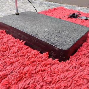 K&ouml;nnte beinhalten: Ein rechteckiger, dunkelgrauer Sockel mit strukturierter Oberfl&auml;che, m&ouml;glicherweise f&uuml;r einen Lichtst&auml;nder, liegt auf einem leuchtend roten, zotteligen Teppich. Ein silberner Pfosten erstreckt sich von der Basis. Stromkabel sind sichtbar.