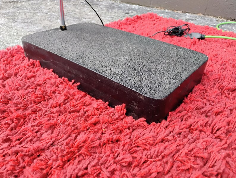 K&ouml;nnte beinhalten: Ein rechteckiger, dunkelgrauer Sockel mit strukturierter Oberfl&auml;che, m&ouml;glicherweise f&uuml;r einen Lichtst&auml;nder, liegt auf einem leuchtend roten, zotteligen Teppich. Ein silberner Pfosten erstreckt sich von der Basis. Stromkabel sind sichtbar.