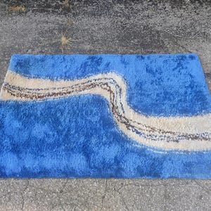 Puede incluir: Una alfombra rectangular de pelo largo con una base azul vibrante y una banda ondulada de color crema que atraviesa el centro en diagonal. La banda crema est&aacute; acentuada por una l&iacute;nea fina, texturizada y de color marr&oacute;n oscuro.