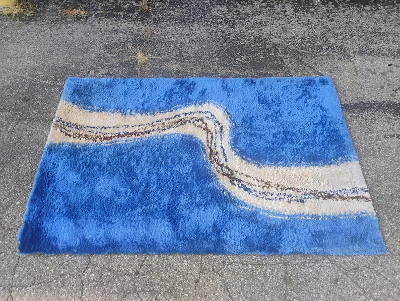 Puede incluir: Una alfombra rectangular de pelo largo con una base azul vibrante y una banda ondulada de color crema que atraviesa el centro en diagonal. La banda crema est&aacute; acentuada por una l&iacute;nea fina, texturizada y de color marr&oacute;n oscuro.