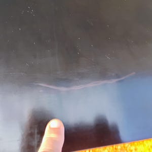 May include: A smooth, black, reflective surface featuring a visible, thin, light-colored scratch or mark running horizontally across the center. The surface appears solid and dark, with subtle light reflections and dust particles visible on the finish.