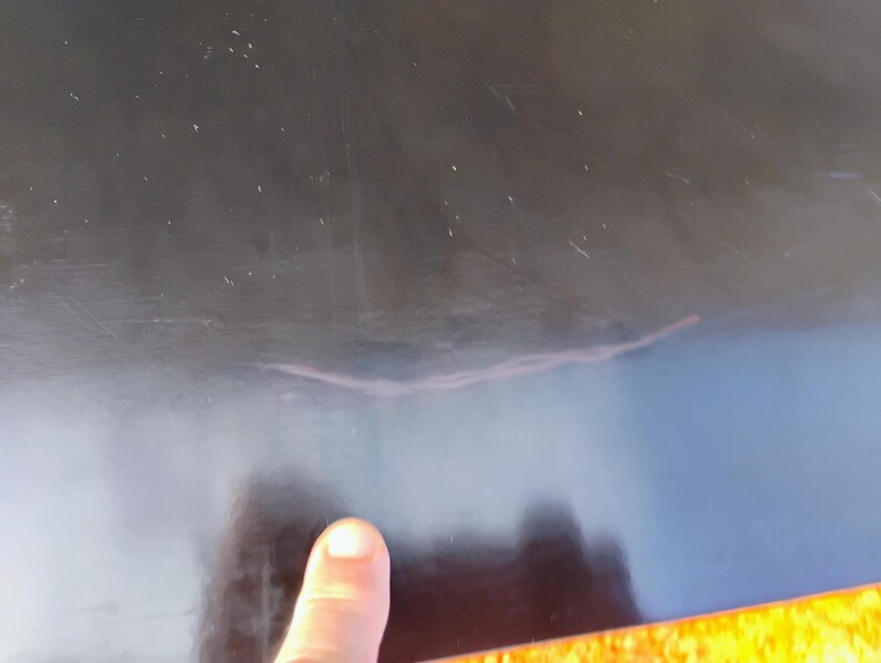 May include: A smooth, black, reflective surface featuring a visible, thin, light-colored scratch or mark running horizontally across the center. The surface appears solid and dark, with subtle light reflections and dust particles visible on the finish.