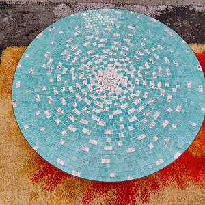 Op de afbeelding: Een ronde turquoise moza&iuml;ek tafelblad met witte tegels in een cirkelvormige opstelling. Het tafelblad is gemaakt van kleine vierkante tegels.