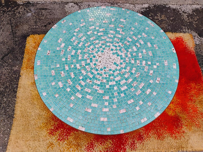 Op de afbeelding: Een ronde turquoise moza&iuml;ek tafelblad met witte tegels in een cirkelvormige opstelling. Het tafelblad is gemaakt van kleine vierkante tegels.