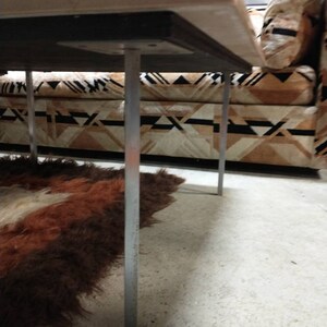 May include: A low-angle view of a rectangular table with a light-colored top and silver legs. The table is positioned over a patterned sofa and a shaggy, brown and white rug. The legs are thin and cylindrical.