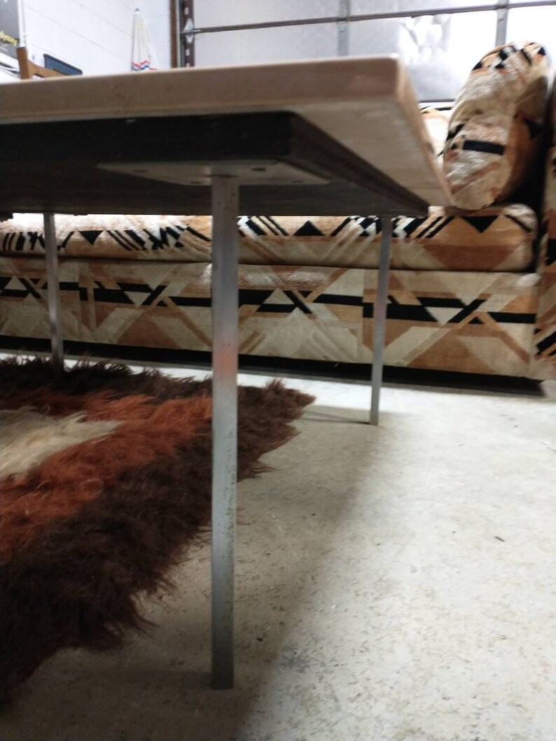 May include: A low-angle view of a rectangular table with a light-colored top and silver legs. The table is positioned over a patterned sofa and a shaggy, brown and white rug. The legs are thin and cylindrical.