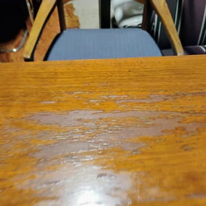 Puede incluir: Primer plano de una mesa de madera con acabado marr&oacute;n claro. La superficie muestra signos de desgaste, con algunos ara&ntilde;azos y marcas de agua.