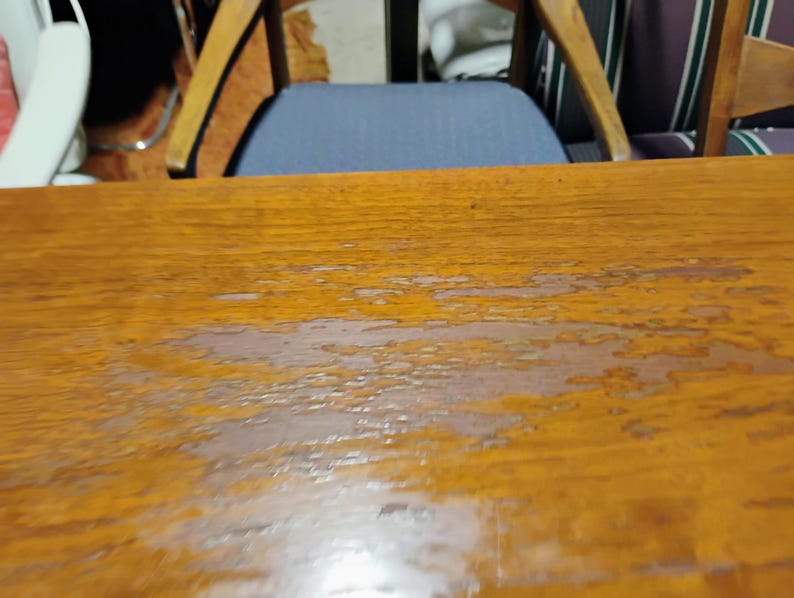 Puede incluir: Primer plano de una mesa de madera con acabado marr&oacute;n claro. La superficie muestra signos de desgaste, con algunos ara&ntilde;azos y marcas de agua.