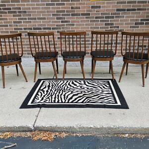 Puede incluir: Cinco sillas de madera con respaldos de varillas y asientos tapizados en negro dispuestas en fila. Cada silla cuenta con cuatro patas c&oacute;nicas y un travesa&ntilde;o superior curvo. Una alfombra rectangular con estampado de cebra blanco y negro.