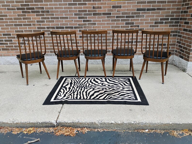 Puede incluir: Cinco sillas de madera con respaldos de varillas y asientos tapizados en negro dispuestas en fila. Cada silla cuenta con cuatro patas c&oacute;nicas y un travesa&ntilde;o superior curvo. Una alfombra rectangular con estampado de cebra blanco y negro.