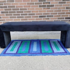 May include: A navy blue upholstered bench with a rectangular shape and rounded corners. The bench is supported by two thick, rectangular legs. A rectangular rug with blue, green, and purple stripes is beneath the bench.