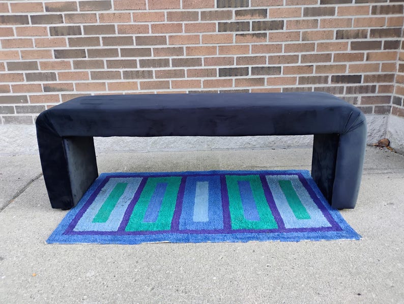 May include: A navy blue upholstered bench with a rectangular shape and rounded corners. The bench is supported by two thick, rectangular legs. A rectangular rug with blue, green, and purple stripes is beneath the bench.