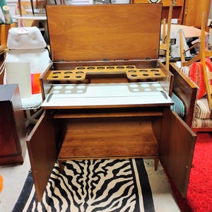 Puede incluir: Un bar de madera vintage con una tapa abatible y un respaldo de espejo. El gabinete tiene dos puertas y un estante en el interior. La parte superior tiene un porta botellas incorporado y una superficie blanca.
