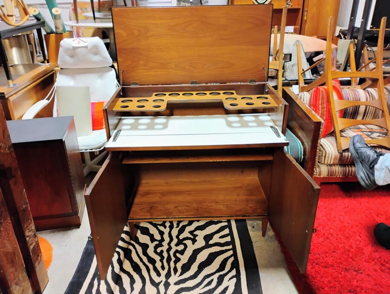 Puede incluir: Un bar de madera vintage con una tapa abatible y un respaldo de espejo. El gabinete tiene dos puertas y un estante en el interior. La parte superior tiene un porta botellas incorporado y una superficie blanca.