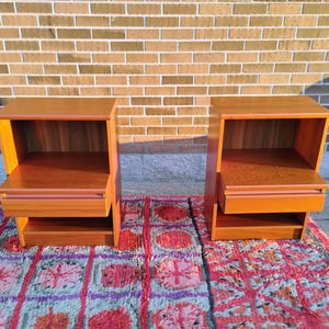 May include: Two matching wooden nightstands with a warm, reddish-brown finish. Each nightstand has an open shelf, a drawer, and a lower shelf. They are placed on a colorful patterned rug in front of a brick wall.