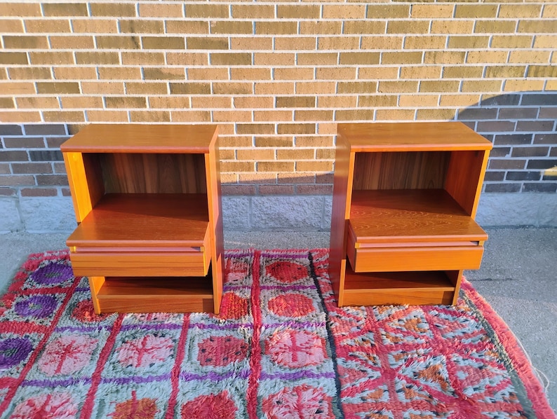 May include: Two matching wooden nightstands with a warm, reddish-brown finish. Each nightstand has an open shelf, a drawer, and a lower shelf. They are placed on a colorful patterned rug in front of a brick wall.