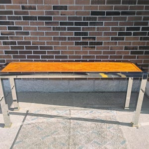 K&ouml;nnte beinhalten: Ein rechteckiger Couchtisch mit polierter Holzplatte und einem verchromten Metallrahmen. Der Tisch hat einen dunklen Rand um das Holz und goldfarbene F&uuml;&szlig;e. Der Tisch ist etwa 122 cm lang.