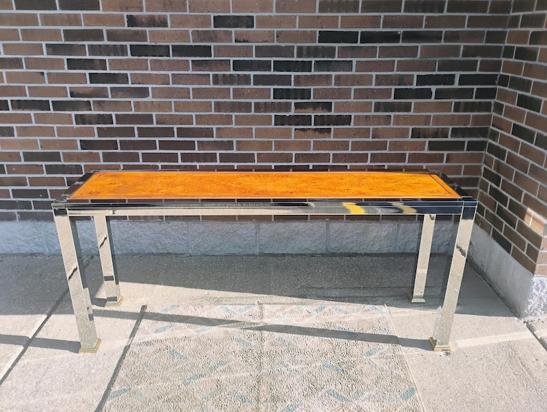 K&ouml;nnte beinhalten: Ein rechteckiger Couchtisch mit polierter Holzplatte und einem verchromten Metallrahmen. Der Tisch hat einen dunklen Rand um das Holz und goldfarbene F&uuml;&szlig;e. Der Tisch ist etwa 122 cm lang.