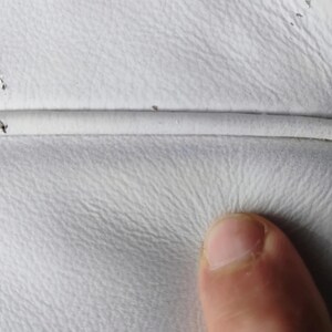 May include: Close-up of white leather upholstery with visible texture and a seam. The image shows minor imperfections and a finger touching the surface, highlighting the material's tactile quality.