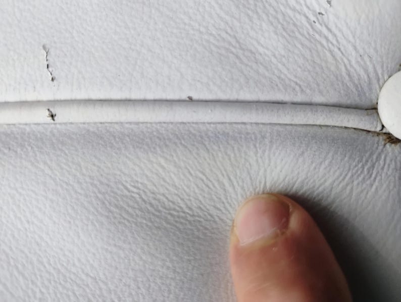 May include: Close-up of white leather upholstery with visible texture and a seam. The image shows minor imperfections and a finger touching the surface, highlighting the material's tactile quality.