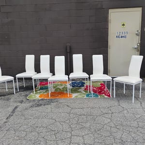 May include: Six white dining chairs with a minimalist design. The chairs have a white seat and backrest, supported by a thin, white metal frame. They are arranged on a colorful floral patterned rug.