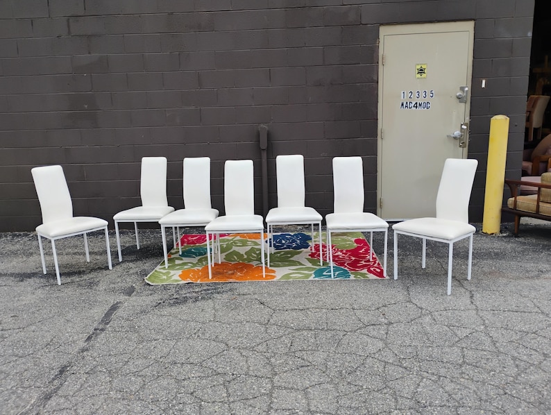 May include: Six white dining chairs with a minimalist design. The chairs have a white seat and backrest, supported by a thin, white metal frame. They are arranged on a colorful floral patterned rug.