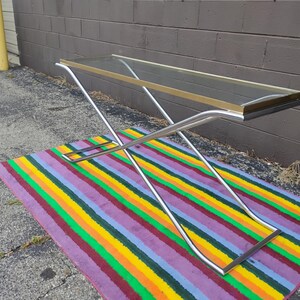 May include: A rectangular console table featuring a clear glass top framed in gold-tone metal. The base is constructed from two intersecting chrome-finished metal tubes that form an X-shape, providing a stable support structure for the tabletop.