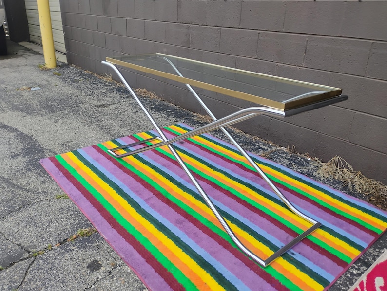 May include: A rectangular console table featuring a clear glass top framed in gold-tone metal. The base is constructed from two intersecting chrome-finished metal tubes that form an X-shape, providing a stable support structure for the tabletop.