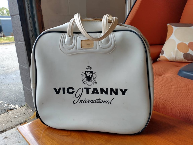 Pode incluir: Bolsa de viagem de couro branco com detalhes pretos e um logotipo preto que diz "VIC TANNY International".