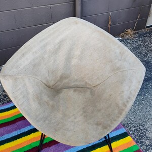 May include: A diamond-shaped chair featuring a beige, textured fabric seat with a visible weave. The chair is supported by thin, black metal legs that extend outward from the base, providing a stable foundation for the upholstered seating area.