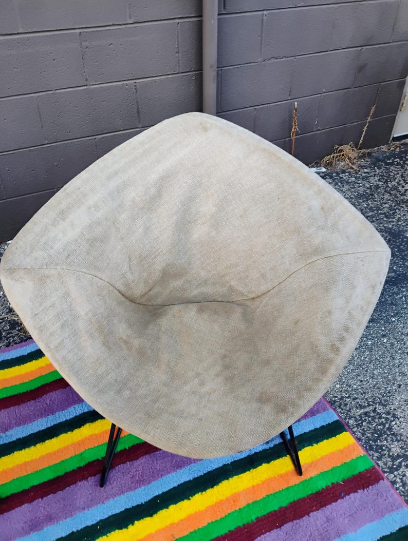May include: A diamond-shaped chair featuring a beige, textured fabric seat with a visible weave. The chair is supported by thin, black metal legs that extend outward from the base, providing a stable foundation for the upholstered seating area.