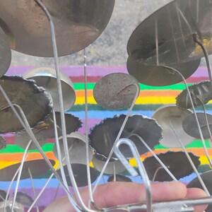May include: A metal sculpture featuring numerous circular discs of varying sizes attached to thin, radiating wire stems. The discs have a metallic, reflective finish with some showing dark, textured edges, all connected to a central metal frame.