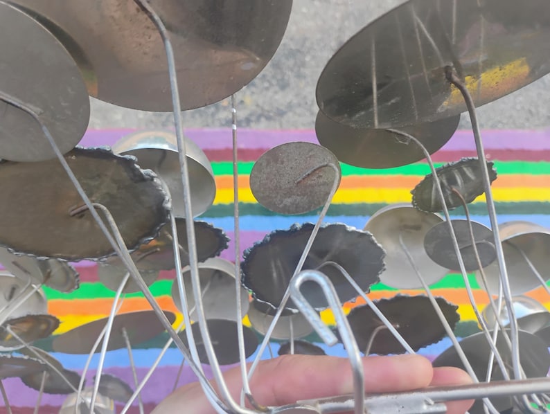 May include: A metal sculpture featuring numerous circular discs of varying sizes attached to thin, radiating wire stems. The discs have a metallic, reflective finish with some showing dark, textured edges, all connected to a central metal frame.